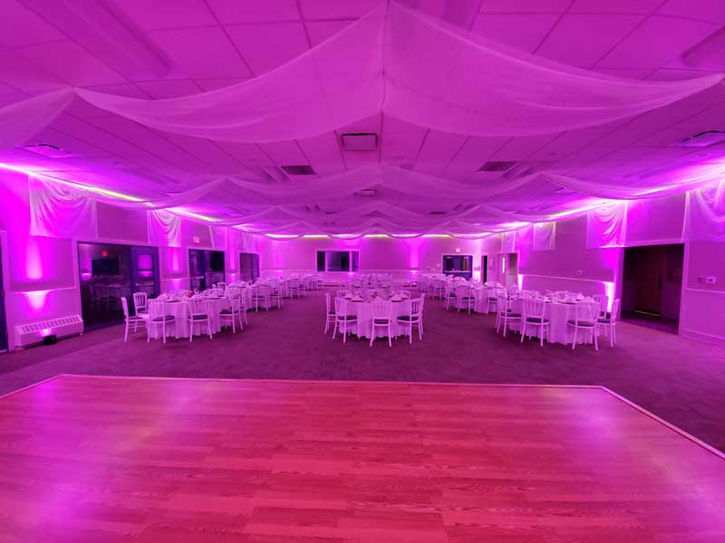 Dining room shown in all pink lighting.  3 rows of table seating 5, 4 table deep for a total of 12 tables.  Curtains drape ceiling and walls, dance floor is set up in front of tables.